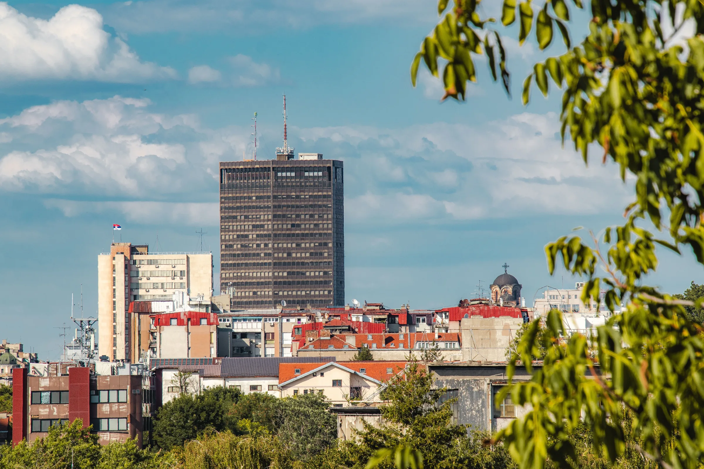 Skyline einer serbischen Stadt: ein Hochhaus über roten Ziegeldächern und einer Kirchenkuppel, von Laub eingerahmt